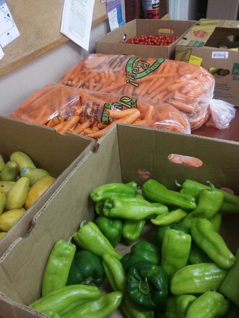 A box of green peppers, A box of pears, a box of cherry tomatoes, and two 50lb bags of carrots set out on a table.